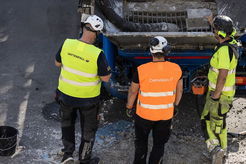 Three construction workers in high-visibility clothing stand behind a concrete mixer truck, coordinating the handling of freshly poured cement on a worksite. Three construction workers in high-visibility clothing stand behind a concrete mixer truck, coordinating the handling of freshly poured cement on a worksite.