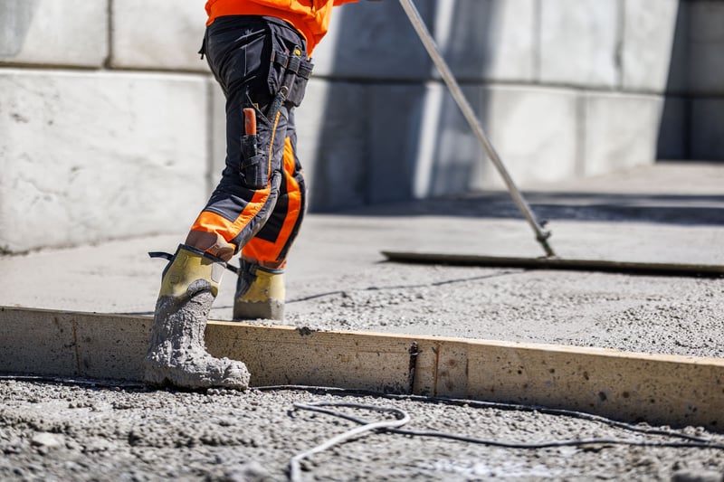 Close-up of a construction worker in high-visibility clothing leveling freshly poured concrete with a screed tool, boots coated in wet cement at a building site. Close-up of a construction worker in high-visibility clothing leveling freshly poured concrete with a screed tool, boots coated in wet cement at a building site.