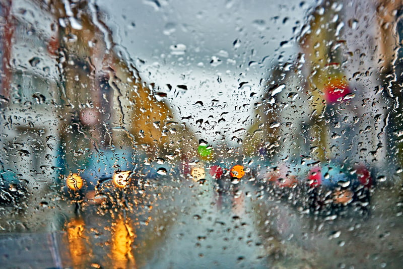 Raindrops on a window with a blurred city street, vehicles and lights visible in the background during wet weather.