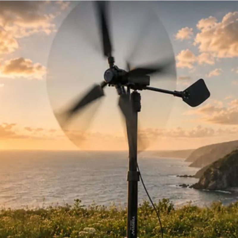 A small wind turbine spinning on a coastal cliff overlooking the sea at sunset, with a rugged shoreline visible in the background.