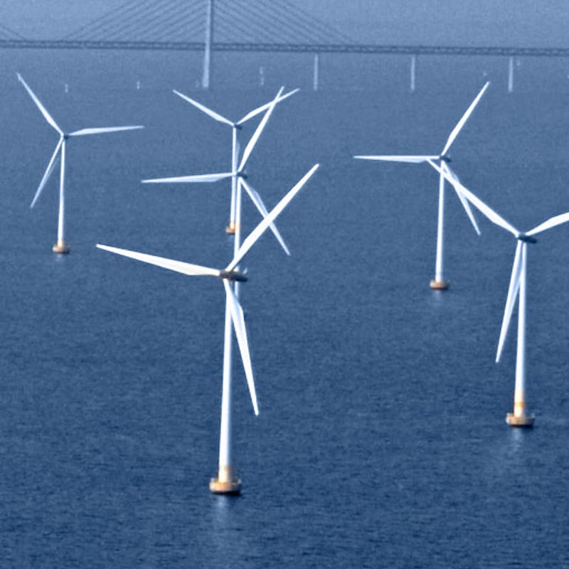 Several offshore wind turbines standing in the sea, viewed from above, with a bridge visible in the background.
