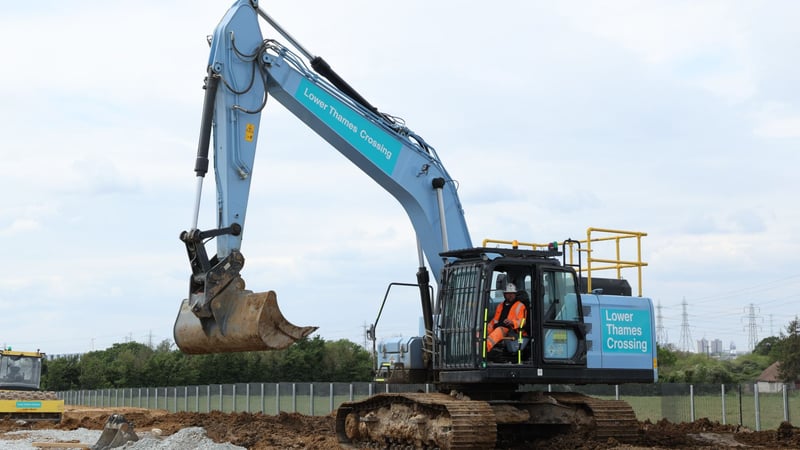 A “Lower Thames Crossing” branded excavator at a construction site, with a worker in high-visibility clothing seated in the cab. A “Lower Thames Crossing” branded excavator at a construction site, with a worker in high-visibility clothing seated in the cab.