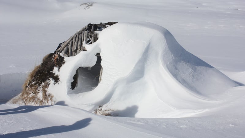 Snow-covered traditional hut partially buried under deep snow in an open winter landscape.