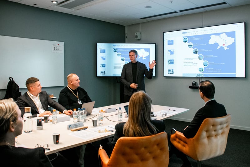 Presenter speaking to participants seated around a conference table, with a presentation showing a map of Ukraine displayed on two screens.