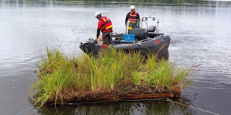 Floating island with vegetation in a river, positioned in front of a small work boat. Photo: Maidul Choudhury.