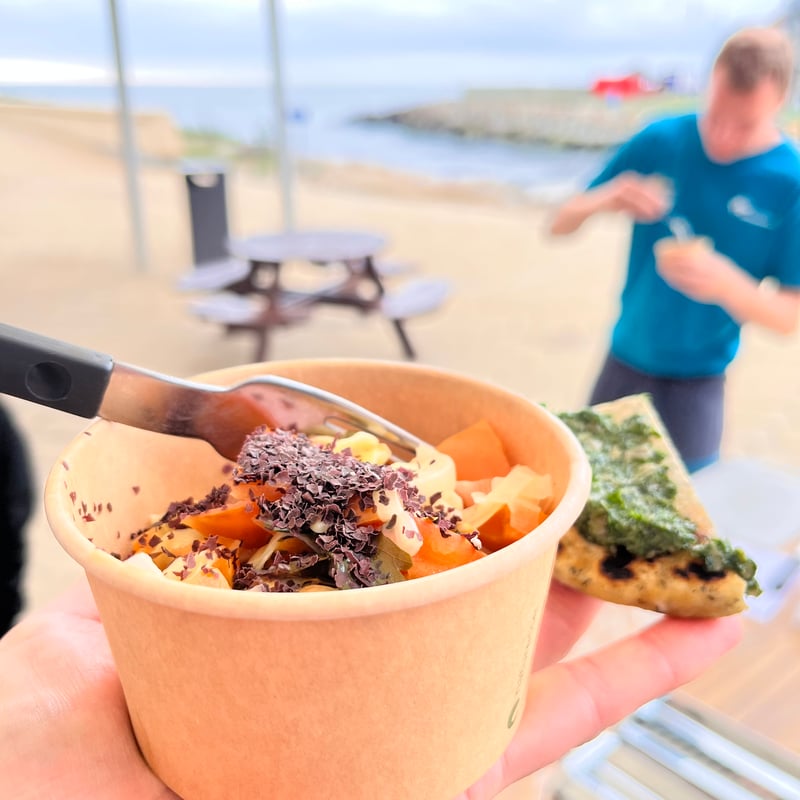 In the foreground, a bowl of food harvested from the sea resting on the palm of a hand - in the background, a man eating such a bowl.