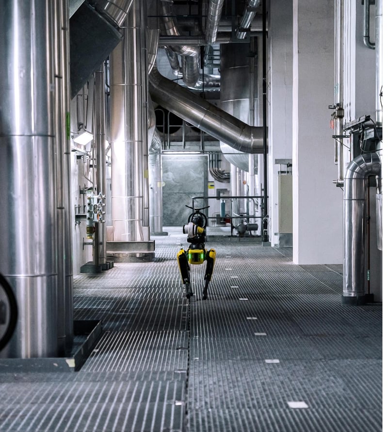 The robot dog Spot walking along a metal-grated floor inside an industrial facility with large pipes, ducts, and stainless-steel equipment.