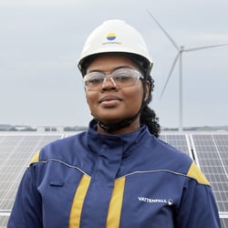 Eine Mitarbeiterin von Vattenfall in Schutzkleidung und mit Schutzhelm steht vor Sonnenkollektoren, im Hintergrund ist eine Windkraftanlage zu sehen.