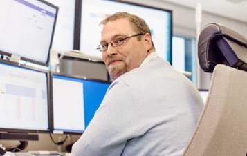 Mitarbeiter vor Monitoren sitzend in Kamera blickend Mitarbeiter vor Monitoren sitzend in Kamera blickend