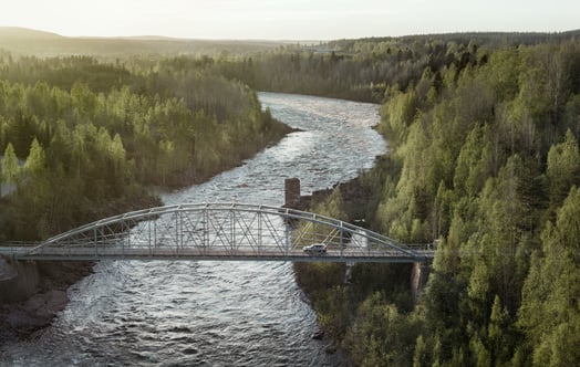 Ein Auto überquert eine von Wald umgebene Stahlbrücke.
