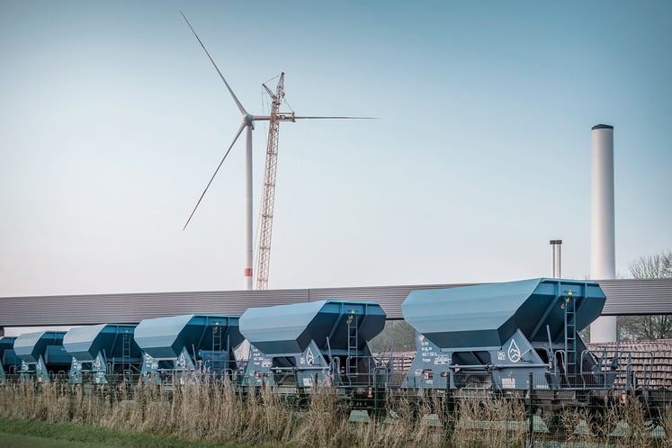 Blaue Güterwagen auf Bahngleisen mit einer Windkraftanlage, einem Kran und Industrieschornsteinen im Hintergrund. Blaue Güterwagen auf Bahngleisen mit einer Windkraftanlage, einem Kran und Industrieschornsteinen im Hintergrund.