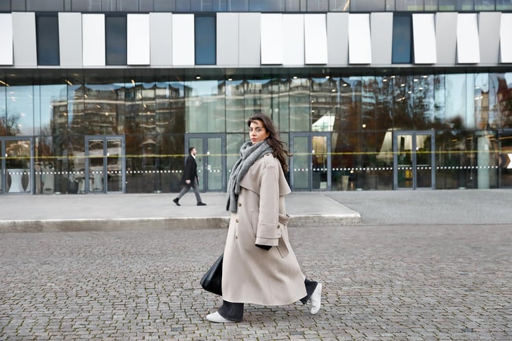 Eine Frau in langem Mantel auf einer gepflasterten Fläche, vor einem modernen Glasgebäude.