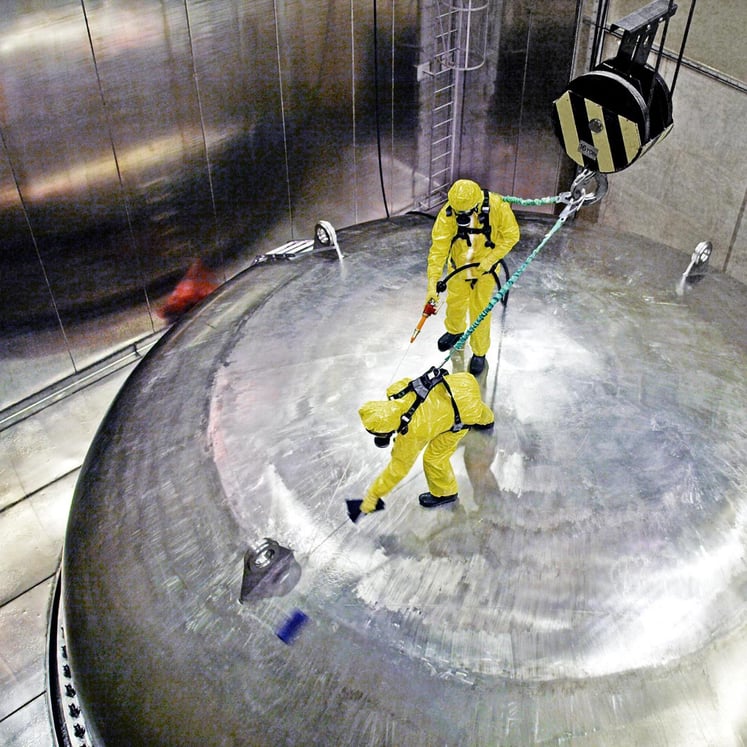 Zwei Personen in gelben Schutzanzügen reinigen einen großen Metalltank in einem Kernkraftwerk. Zwei Personen in gelben Schutzanzügen reinigen einen großen Metalltank in einem Kernkraftwerk.