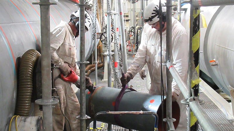 Zwei Mitarbeiter in Schutzkleidung auf der Vorwärmerbühne zwischen zwei großen Rohren stehend, bearbeiten ein Rohr