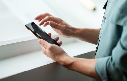 Hands holding and using a smartphone near a window.