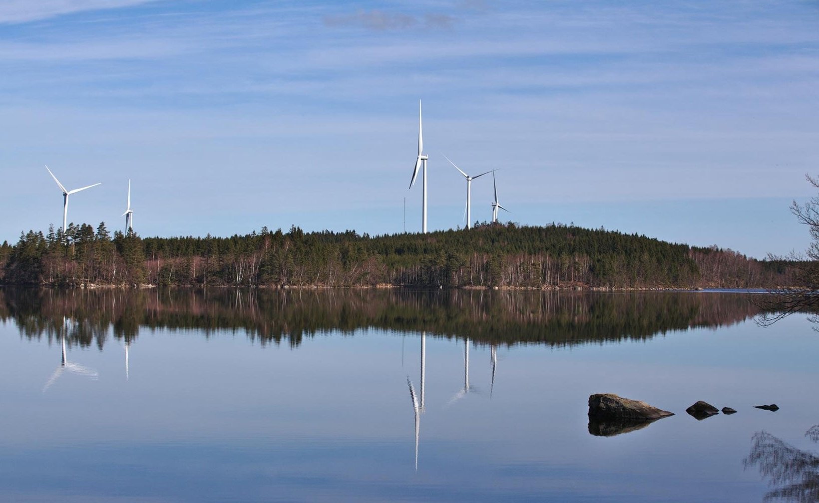 Tuulivoimalat metsäisen rantaviivan varrella heijastuvat tyynessä järvessä kirkkaan taivaan alla.