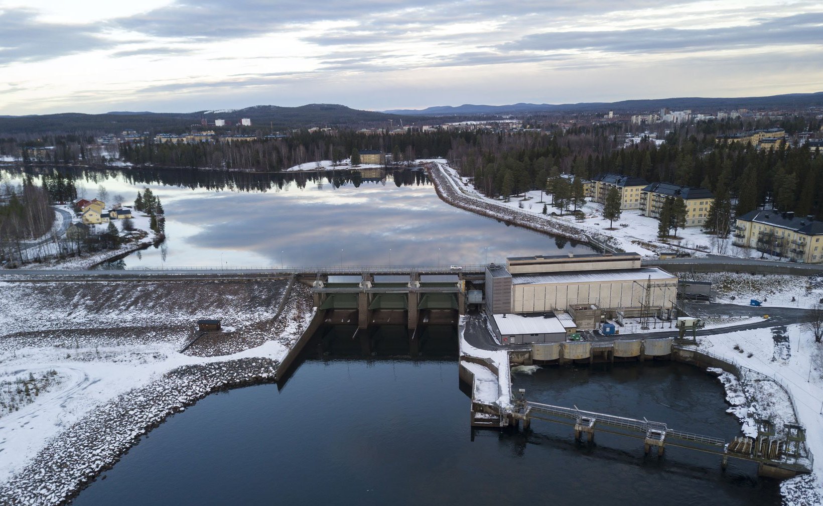 Ilmakuva vesivoimalaitoksesta ja padosta talvisessa maisemassa, jossa joki ja tekojärvi ympäröivät metsää ja lähellä sijaitsevia rakennuksia.