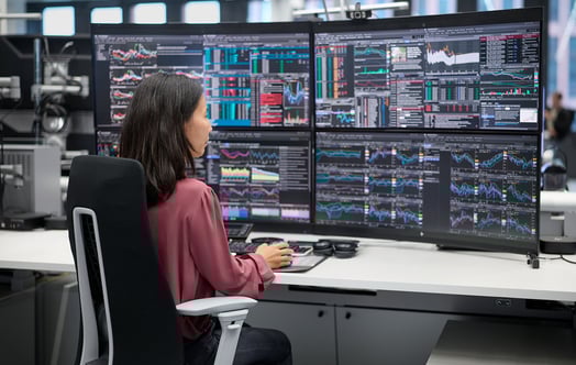 Nainen työskentelee työpöydän ääressä, jossa on neljä suurta näyttöä, joilla näkyy pörssikurssikaavioita ja -tietoja.