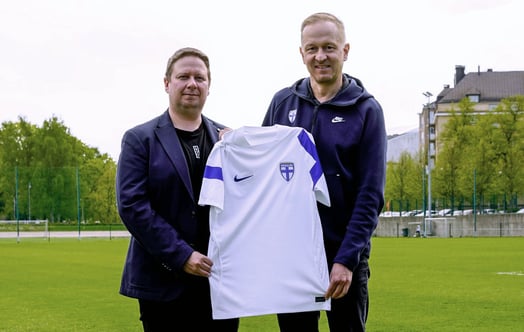 Kaksi henkilöä seisoo jalkapallokentällä pitäen käsissään valkoista Suomen maajoukkuepaitaa sinisin yksityiskohdin, taustalla maali ja rakennuksia.