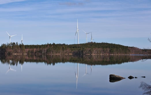 Tuulivoimalat metsäisen rantaviivan varrella heijastuvat tyynessä järvessä kirkkaan taivaan alla.