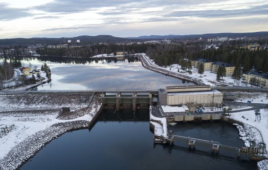 Ilmakuva vesivoimalaitoksesta ja padosta talvisessa maisemassa, jossa joki ja tekojärvi ympäröivät metsää ja lähellä sijaitsevia rakennuksia.