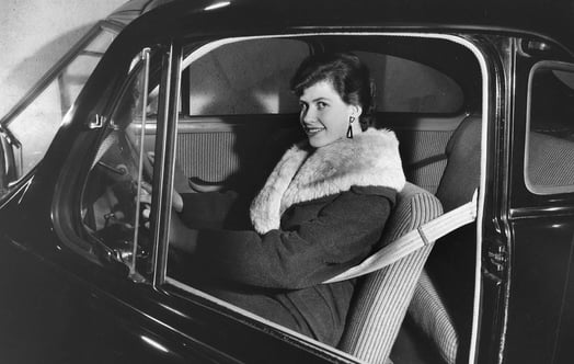 Mustavalkoinen kuva naisesta, joka istuu vanhemman mallin autossa turvavyö kiinnitettynä. Mustavalkoinen kuva naisesta, joka istuu vanhemman mallin autossa turvavyö kiinnitettynä.