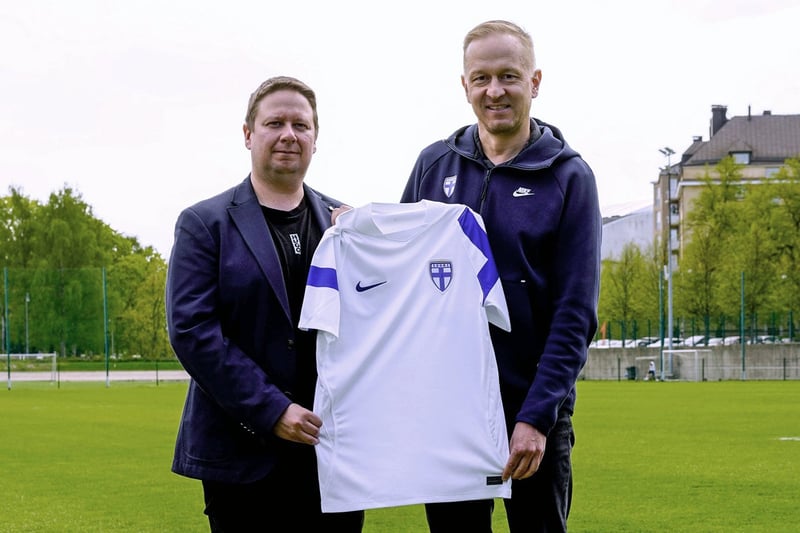 Kaksi henkilöä seisoo jalkapallokentällä pitäen käsissään valkoista Suomen maajoukkuepaitaa sinisin yksityiskohdin, taustalla maali ja rakennuksia.