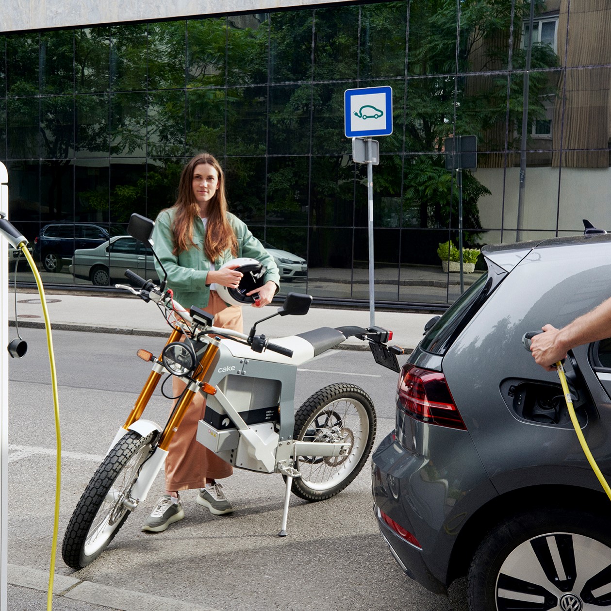 Frau steht hinter einem elektrischem Motorrad, E-Auto wird von Mann geladen