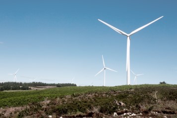 Image of a wind turbine located on a hill, in a green landscape with blue skies above.