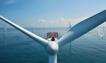 Image zoomed in on a Vattenfall employee, working from the top of a wind turbine located in the sea. More wind turbines and a small boat is in the background.