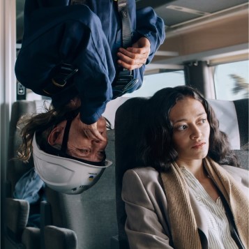 A woman sits in a train and a wind turbine worker is hanging upside down whispering in her ear