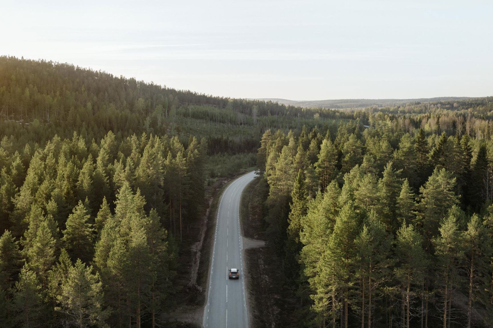 Flygbild över en bil som kör på en slingrande väg genom tät barrskog.