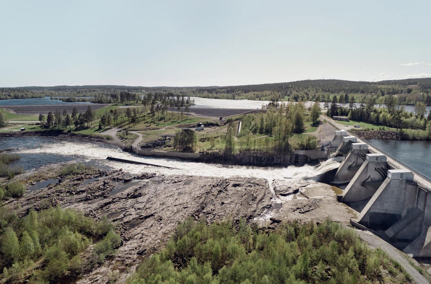 Flygvy över vattenkraftverk med forsande vatten, omgiven av grönskande landskap