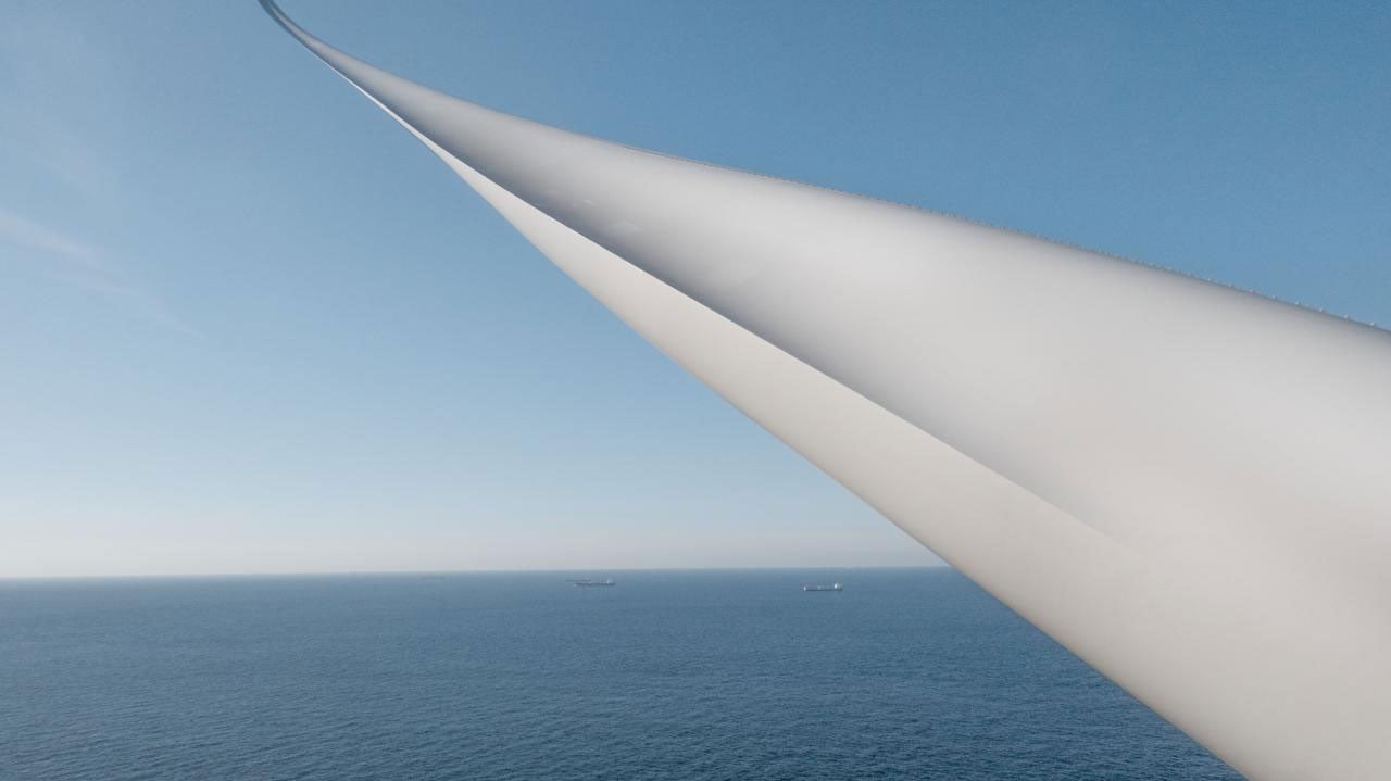 Vindkraftsblad över havet under klar blå himmel, med två fartyg vid horisonten.