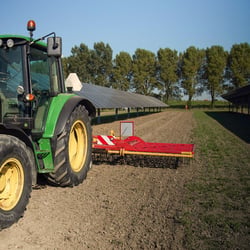 Rader av solpaneler längs en odlad åker, med en traktor som arbetar mellan panelraderna.
