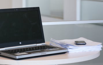 Laptop och en trave papper på ett skrivbord i kontorsmiljö.