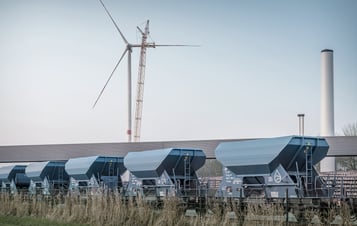 Blå godsvagnar på järnvägsspår med ett vindkraftverk, en kran och industriskorstenar i bakgrunden.