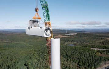Ett maskinhus till ett vindkraftverk lyfts på plats på en turbin, med skogslandskap och andra vindkraftverk i bakgrunden.