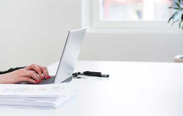 Händer på en bärbar dator bredvid en trave papper
