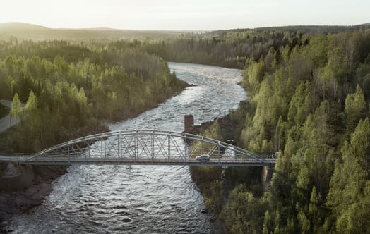 Bil på en stålbro över älv i skogslandskap.