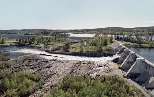 Flygvy över vattenkraftverk med forsande vatten, omgiven av grönskande landskap