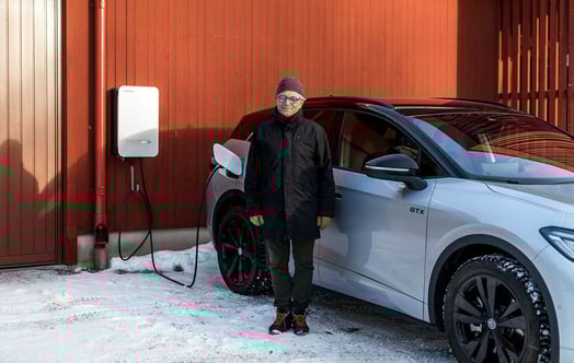 En man står i snön bredvid en elbil som laddas vid en röd träbyggnad.
