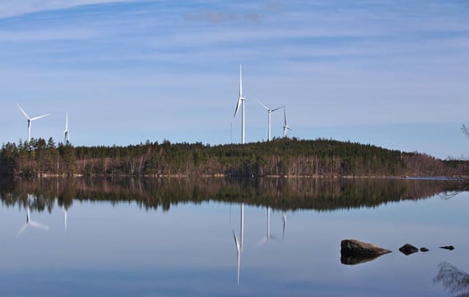 Vindkraftverk på en skogsklädd höjd speglas i en stilla sjö under klar himmel.