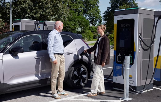 Två personer står vid en elbil intill en laddstation på en solig parkering.