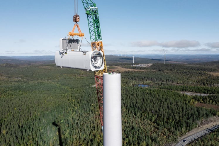 Ett maskinhus till ett vindkraftverk lyfts på plats på en turbin, med skogslandskap och andra vindkraftverk i bakgrunden.