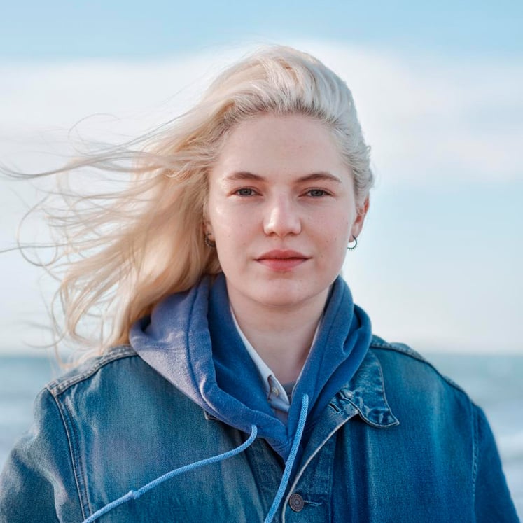 Person med långt blont hår i blå jeansjacka vid havet.