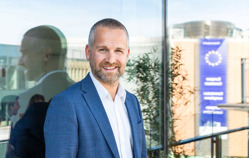 Erik Filipsson, Public and Regulatory Affairs Advisor på Vattenfall, i blå kavaj och vit skjorta står vid en glasvägg med spegling; modern byggnad och banderoll i bakgrunden.