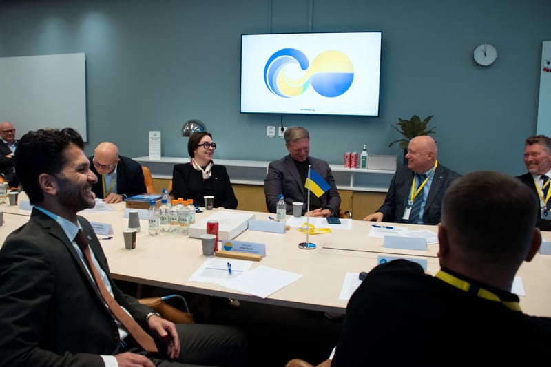 Deltagare sitter runt ett konferensbord under ett möte, med en liten ukrainsk flagga på bordet och en blågul grafik på en skärm.