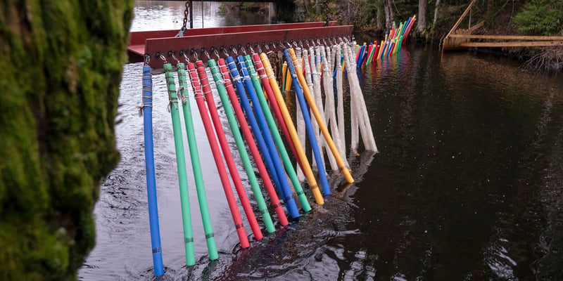 Färgglada plaststavar upphängda i en rad från en röd balk ner i en smal å. Foto: Johan Gunséu. Färgglada plaststavar upphängda i en rad från en röd balk ner i en smal å. Foto: Johan Gunséu.