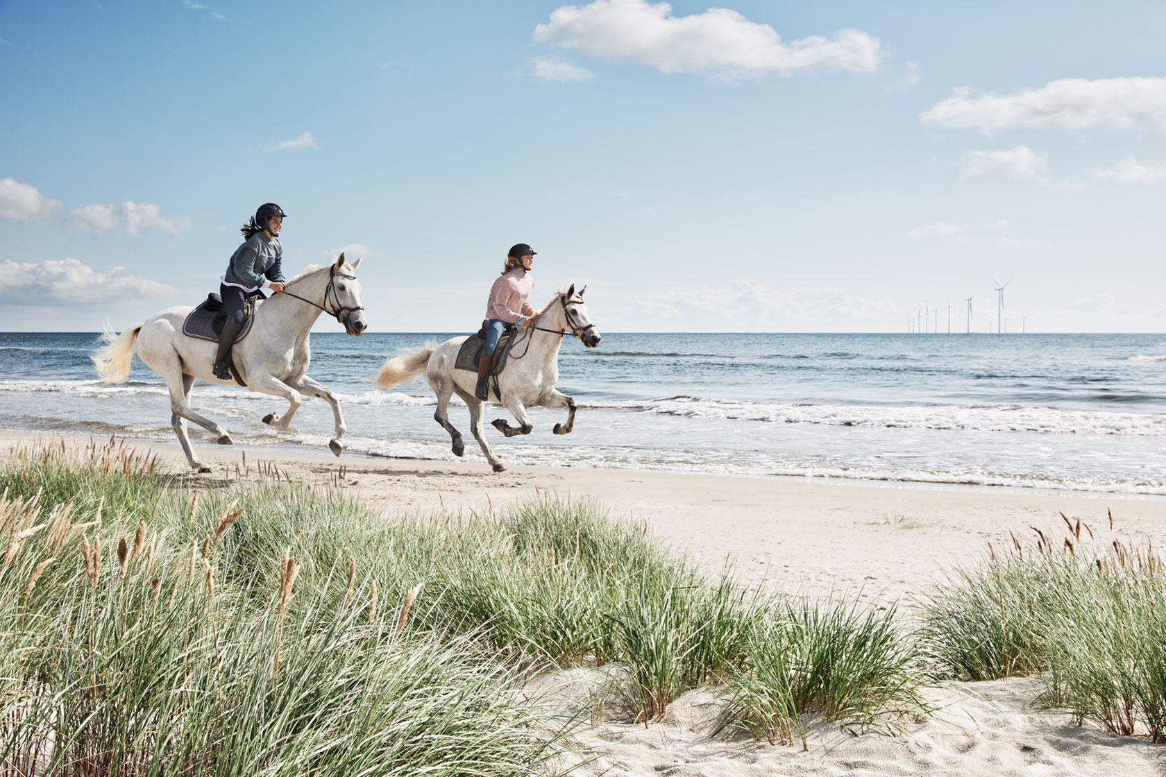 Två ryttare rider sina hästar på en strand med vindkraftverk i bakgrunden